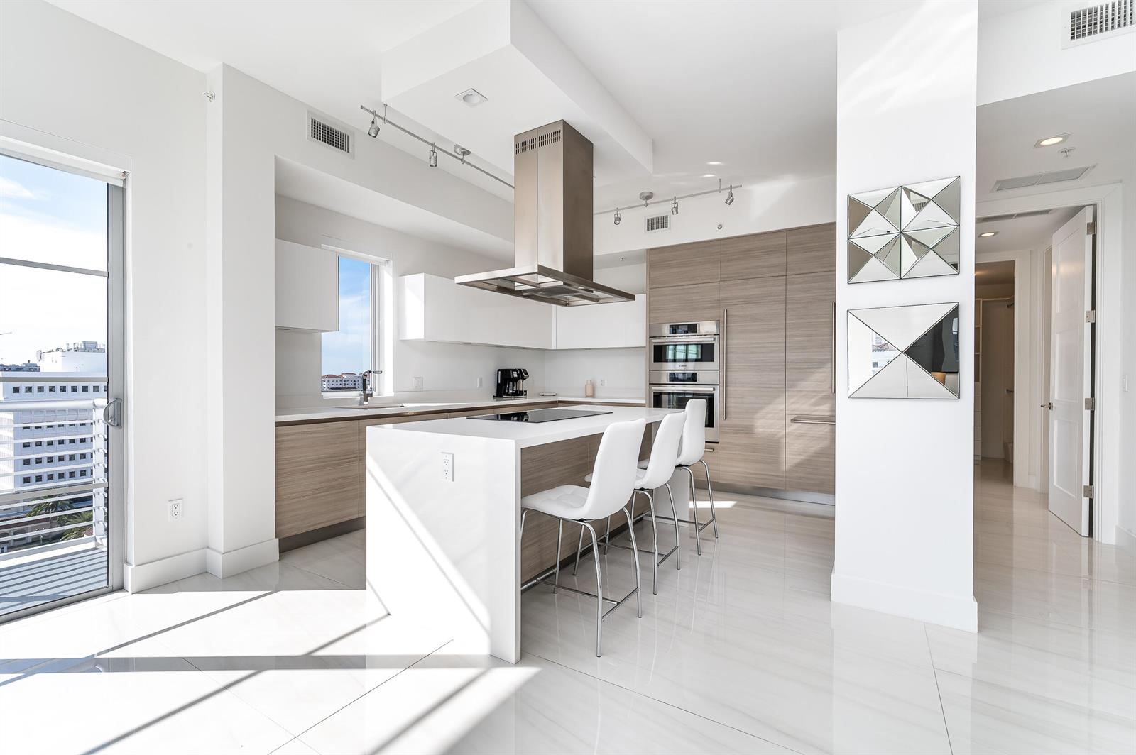 A modern kitchen features a white island with bar stools, wood cabinetry, and stainless appliances — 155 E Boca Raton Road # 1103, Boca Raton FL 33432, BOCA RATON, FL
