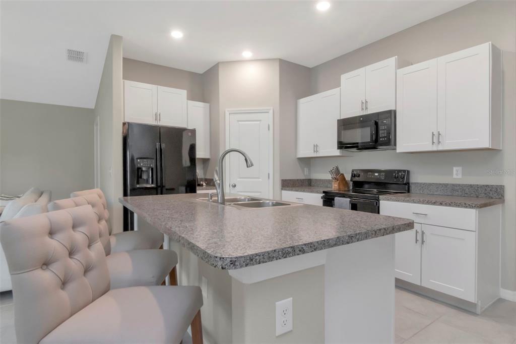 A kitchen with white cabinets, a granite island, black appliances, and gray tufted bar stools — 11527 SAGE CANYON DRIVE, RIVERVIEW, FL