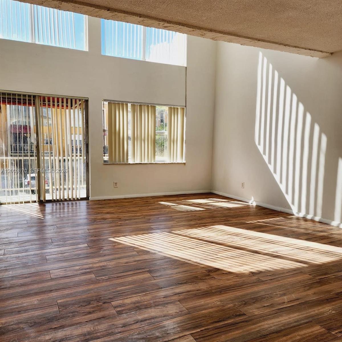 An empty room features high ceilings, wood-look flooring, and large windows with vertical blinds — 18051 NW 68th Ave # K202, Hialeah FL 33015, HIALEAH, FL