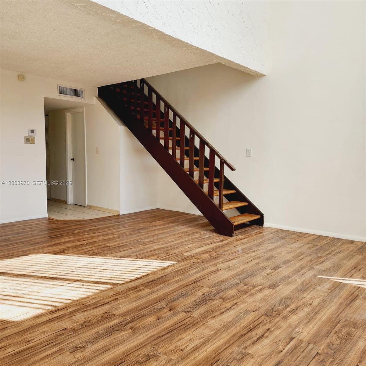 An empty room features wood-look flooring, white walls, and a dark wooden staircase — 18051 NW 68th Ave # K202, Hialeah FL 33015, HIALEAH, FL