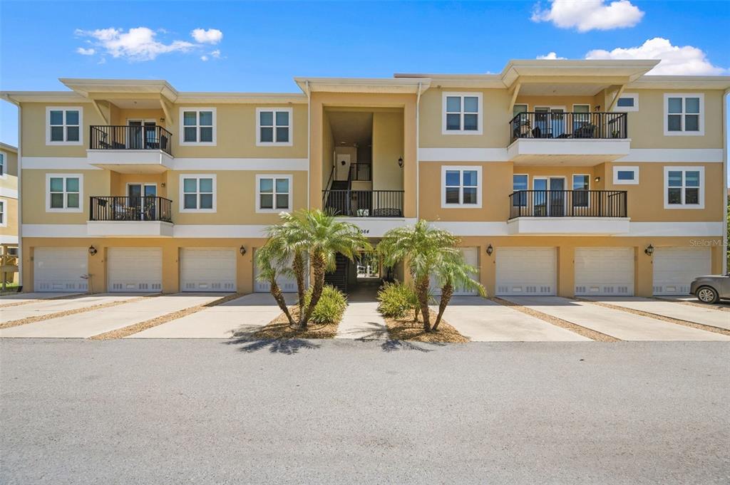A three-story apartment building with tan stucco walls, multiple balconies, and ground-level garage doors — 5064 ROYAL PALMS WAY UNIT 204, NEW PORT RICHEY, FL