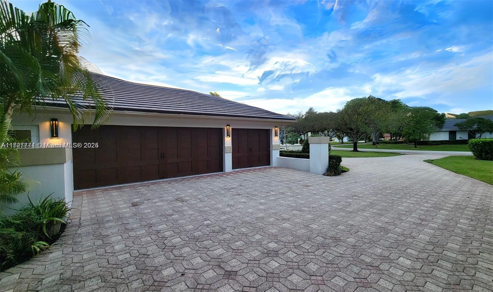 A paved driveway leads to a three-car garage with dark brown doors and exterior lighting — 7700 S Woodridge Dr, Parkland FL 33067, PARKLAND, FL