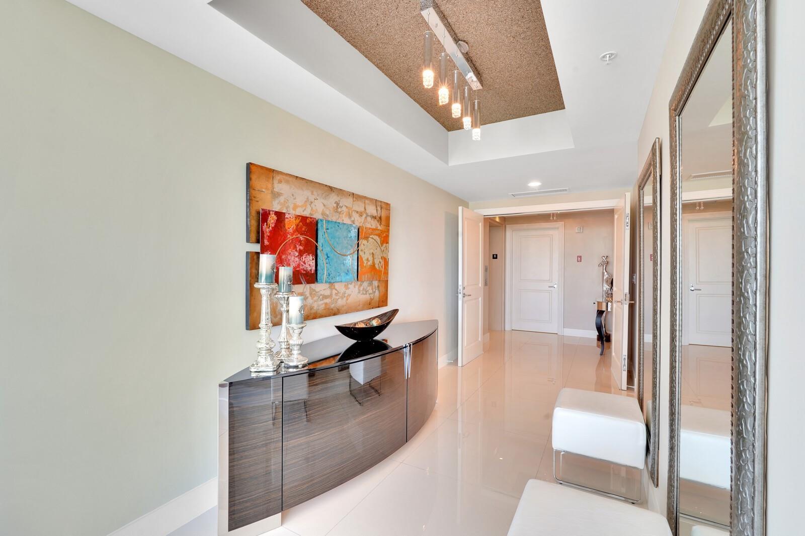 A modern hallway features a curved dark wood console, abstract wall art, and a mirrored wall — 1 N Ocean Boulevard # 607, Pompano Beach FL 33062, POMPANO BEACH, FL