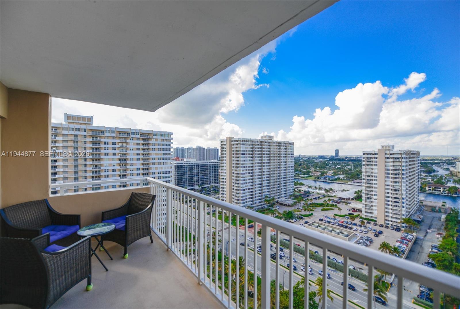 A balcony with wicker chairs and a small table overlooks a city and waterway — 1950 S Ocean Dr # 19E, Hallandale Beach FL 33009, HALLANDALE BEACH, FL