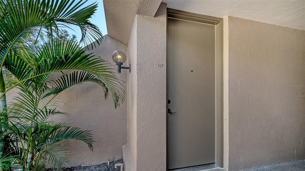 An exterior view of a tan building entrance with a palm tree and unit number 117 — 117 PASS KEY ROAD UNIT 117, SARASOTA, FL
