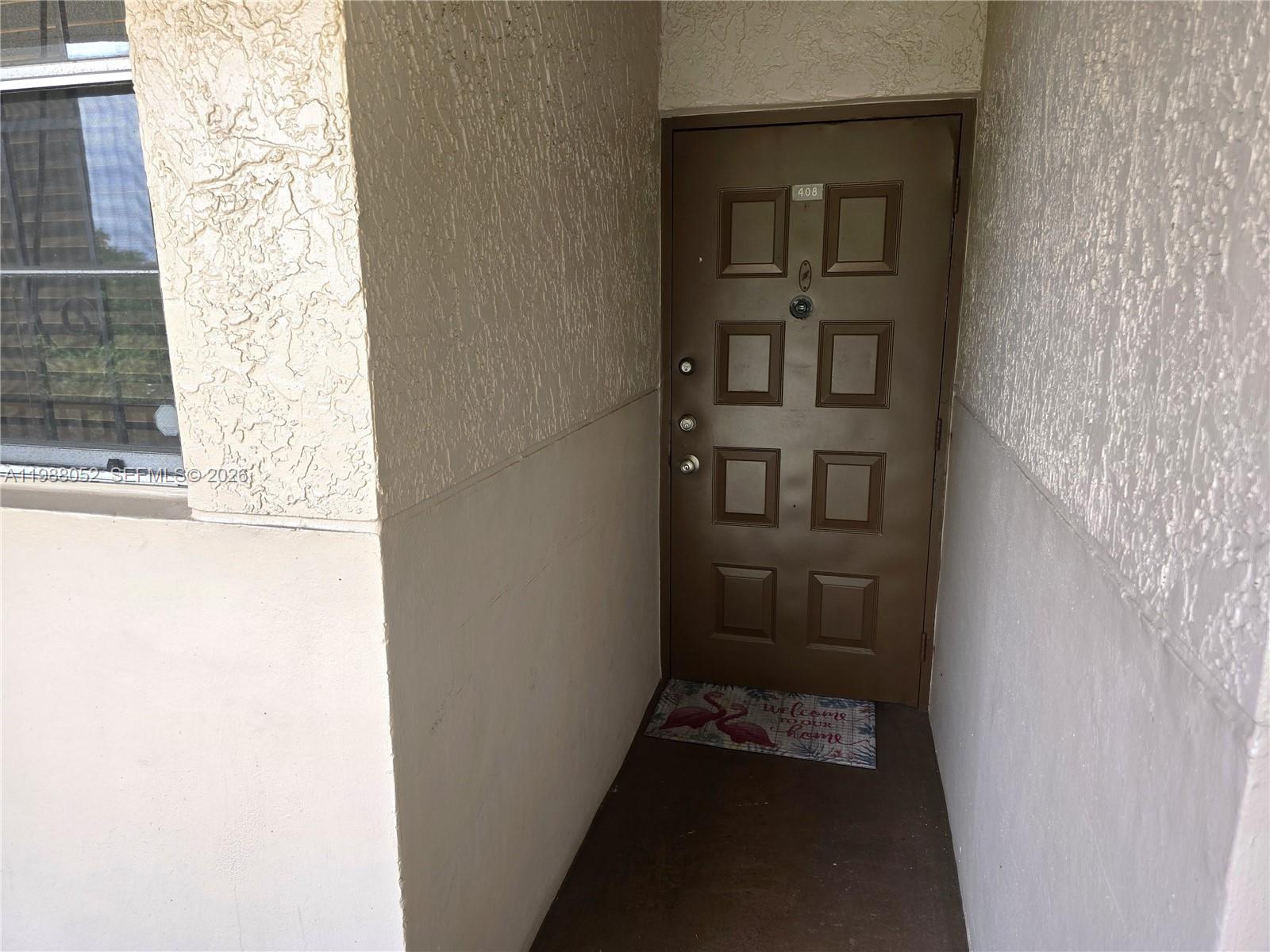 A brown front door with a flamingo welcome mat is set in a stucco alcove — 1811 Sabal Palm Dr # 408, Davie FL 33324, DAVIE, FL