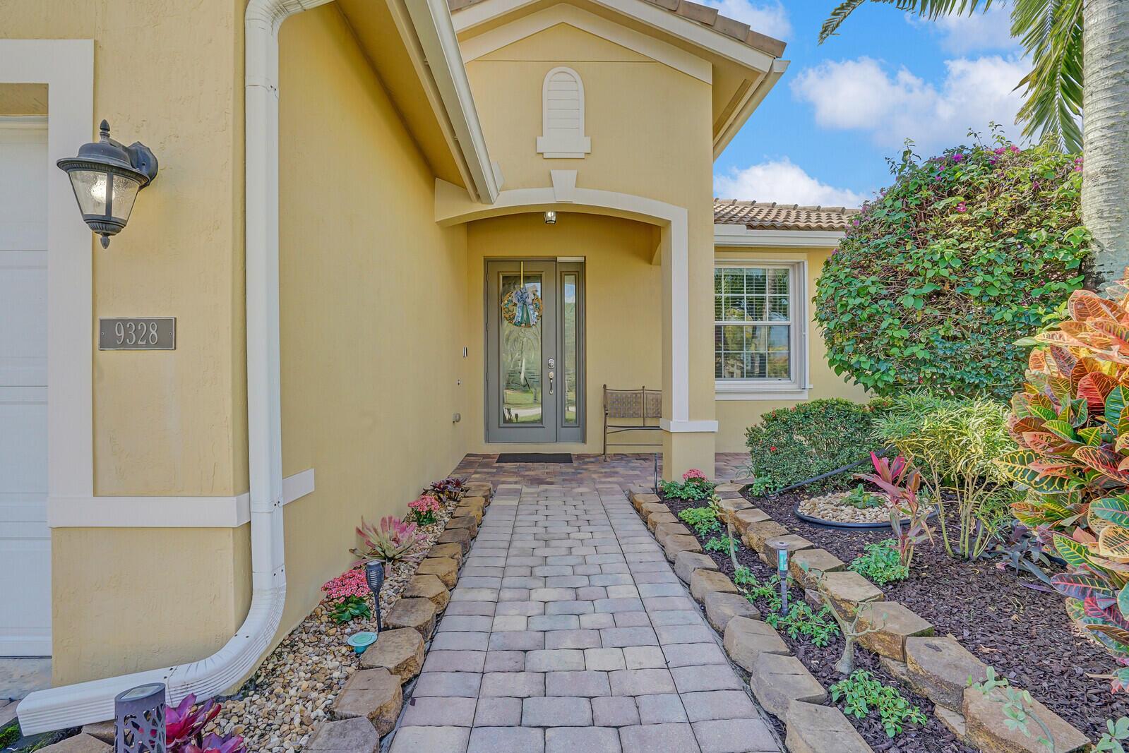 A paved walkway leads to the front entrance of a yellow stucco house with landscaping — 9328 Silent Oak Circle, West Palm Beach FL 33411, WEST PALM BEACH, FL