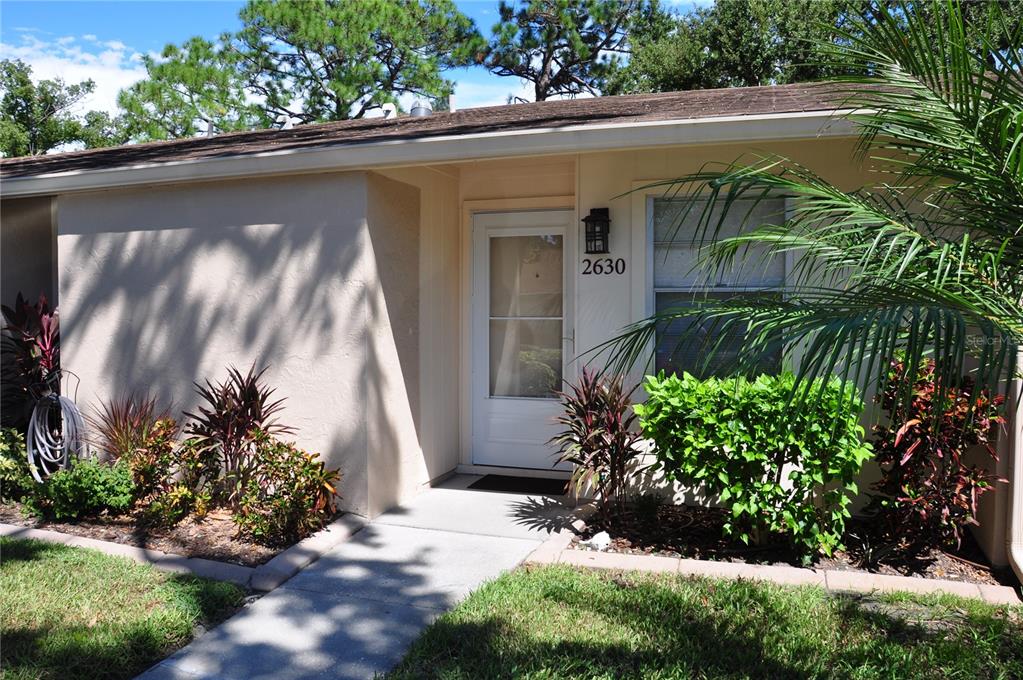The exterior of a single-story home features a tan stucco facade, a white front door, and landscaping — 2630 WOODGATE LANE UNIT G3, SARASOTA, FL
