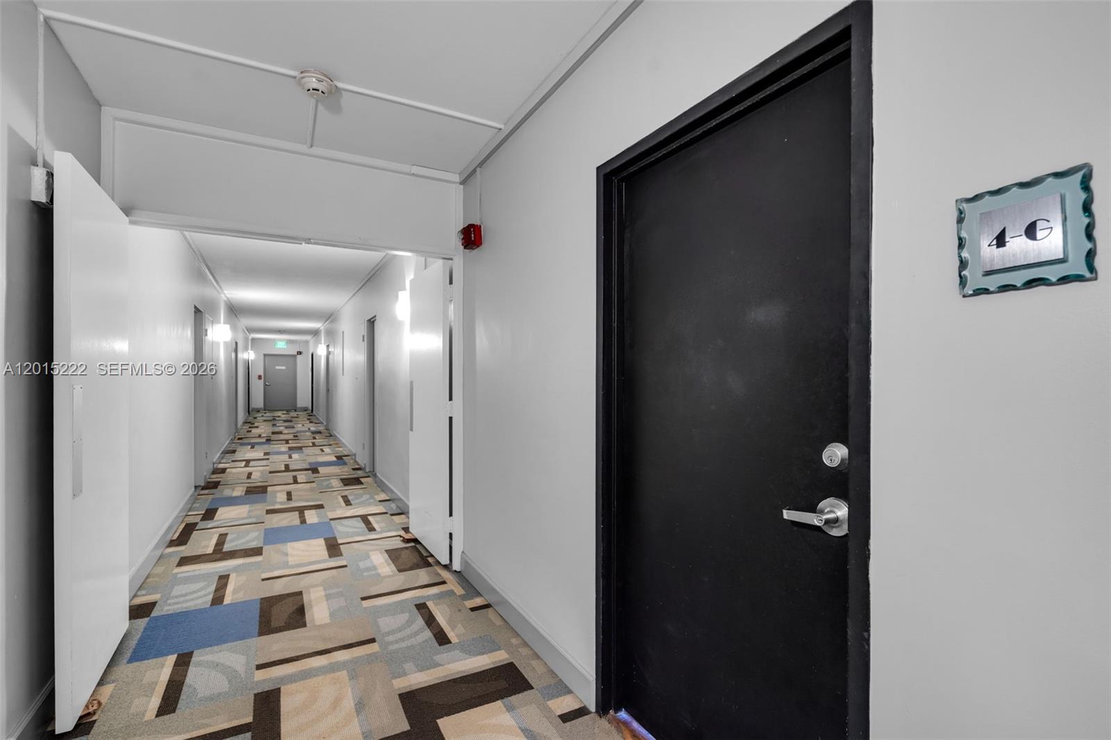 A hallway with patterned carpet leads to a distant door, with a black apartment door nearby — 7441 Wayne Ave # 4G, Miami Beach FL 33141, MIAMI BEACH, FL