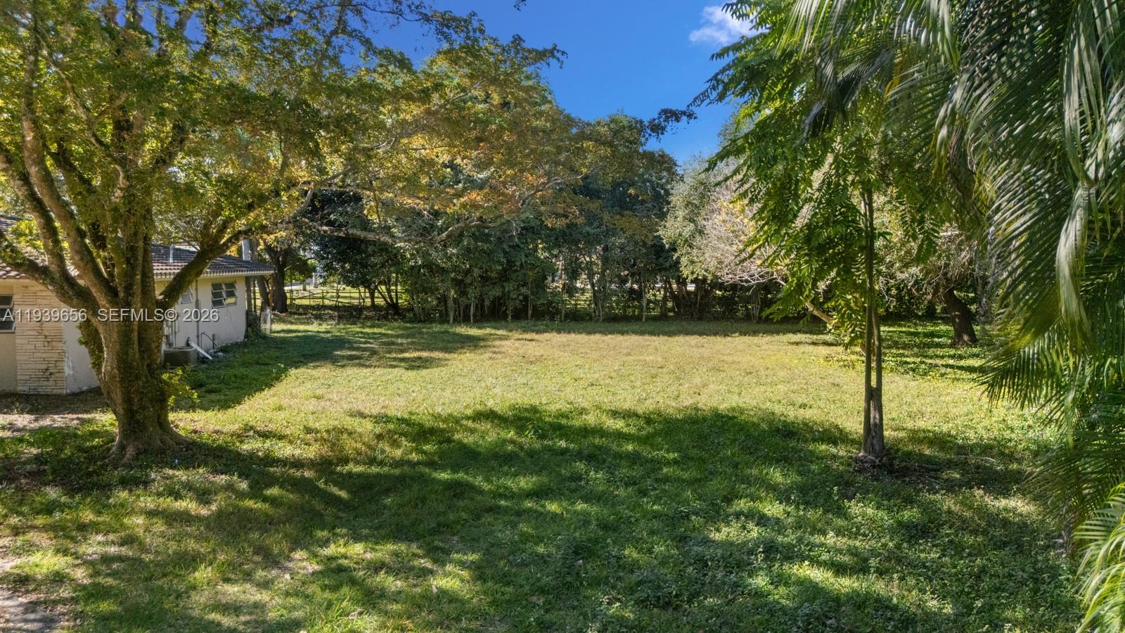A large, grassy backyard features mature trees and a partial view of a house exterior — 13500 SW 68th Ct, Pinecrest FL 33156, PINECREST, FL