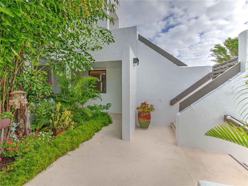 A white stucco building exterior features a covered entryway, lush landscaping, and a wooden staircase — 830 BAYPORT WAY UNIT 830, LONGBOAT KEY, FL