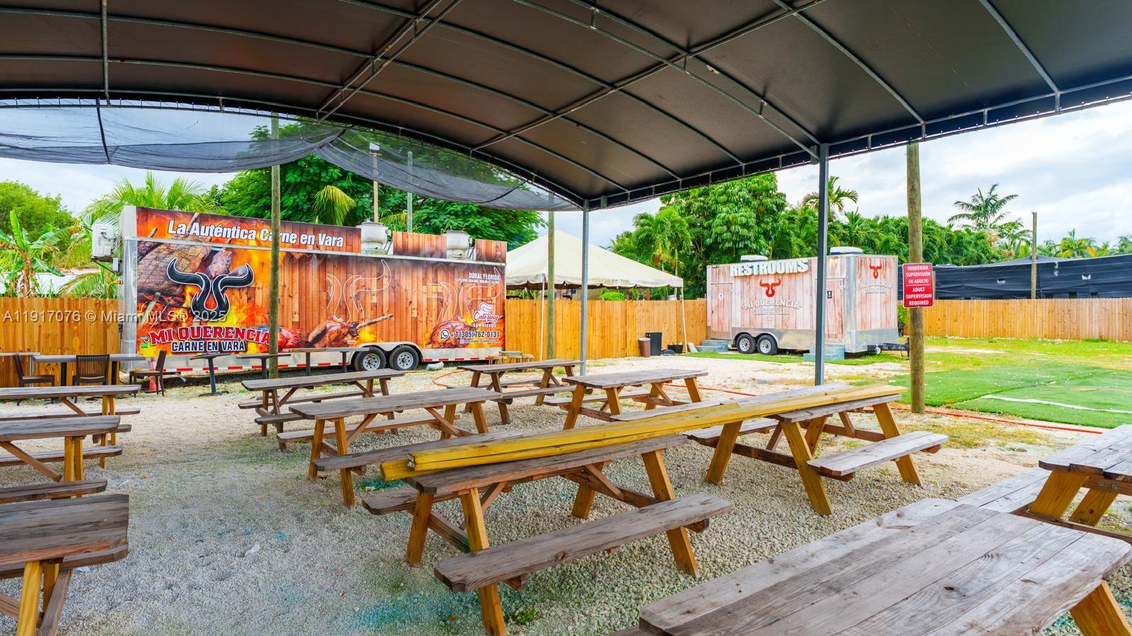 An outdoor dining area features multiple wooden picnic tables under a large canopy near food trailers — 16860 SW 177TH AVE, Miami FL 33187, MIAMI, FL