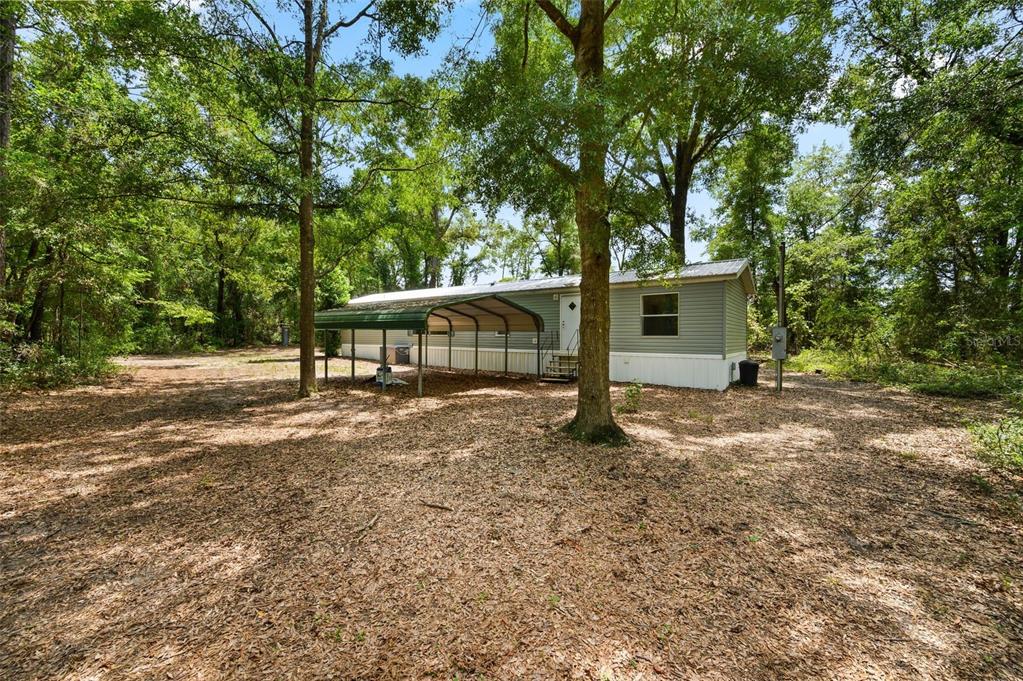 A single-story manufactured home with a metal carport sits on a wooded, leaf-covered lot — 25697 21ST ROAD, O'BRIEN, FL