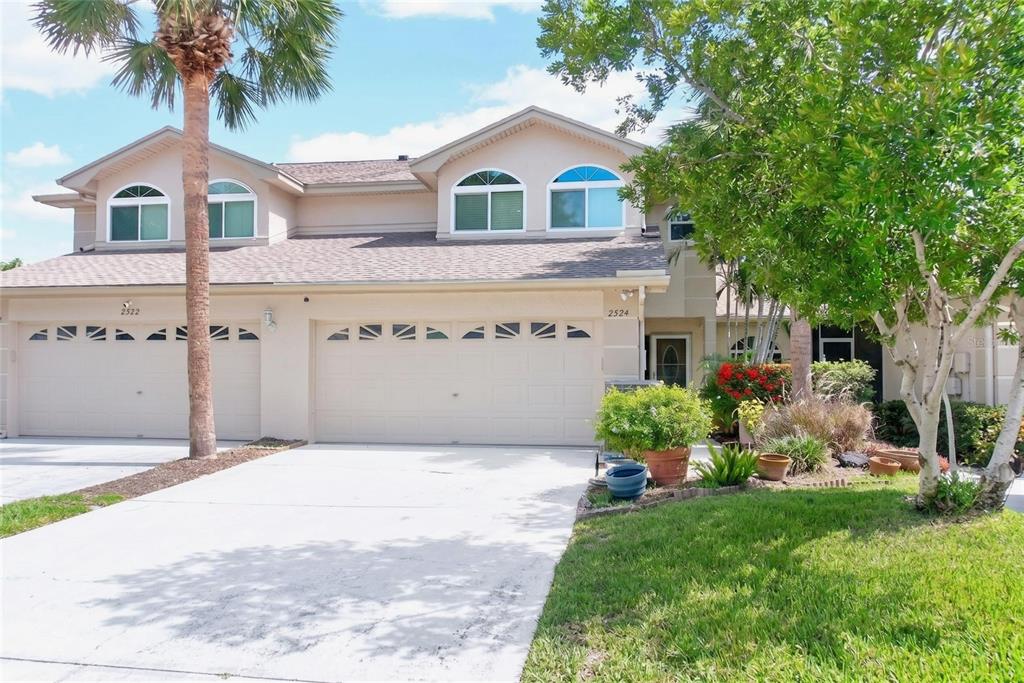 A two-story duplex with beige stucco siding, two garage doors, and a palm tree — 2524 W BROOK LANE, CLEARWATER, FL