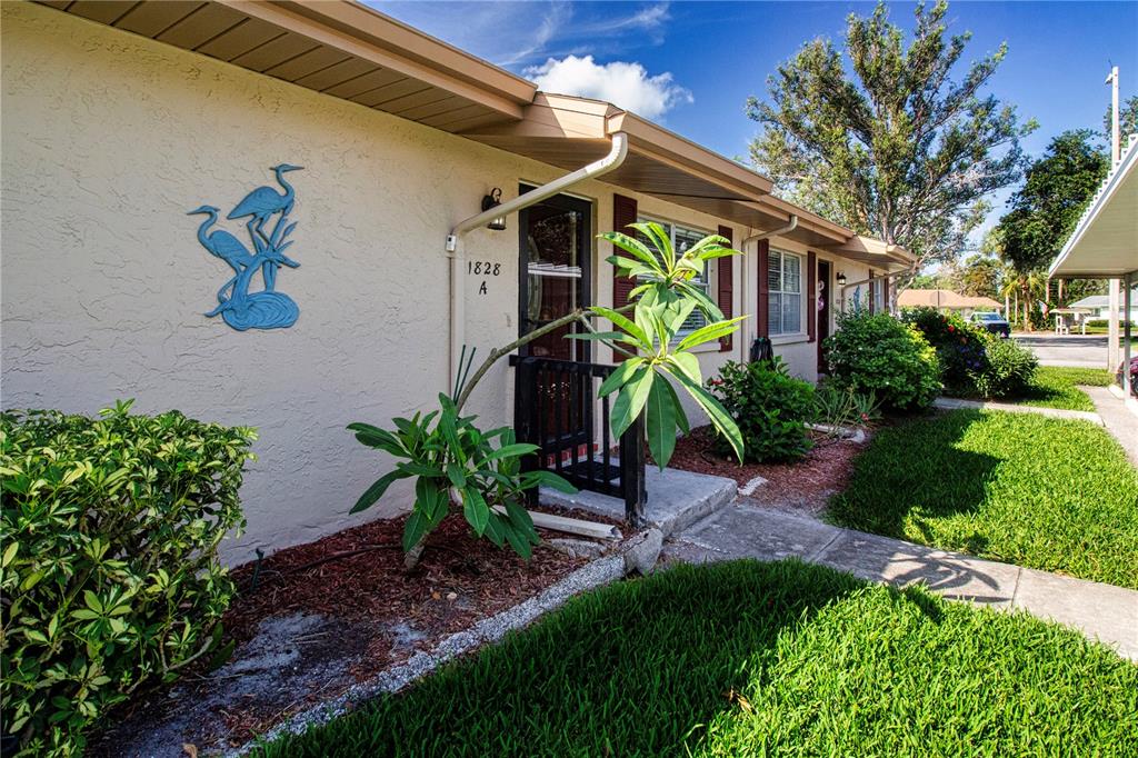 The exterior of a single-story residential unit features a beige stucco wall, a front door, and landscaping — 1828 FOXHUNT DRIVE UNIT A, SUN CITY CENTER, FL