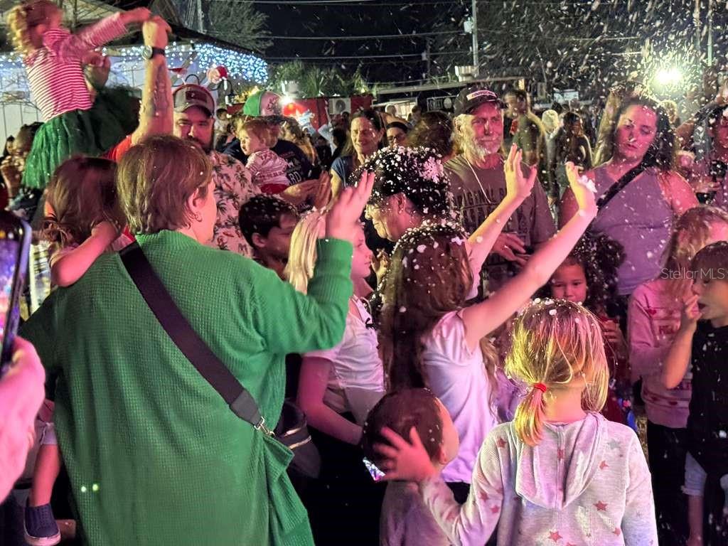 A crowd of children and adults gathers outdoors at night while artificial snow falls — 57TH STREET, DUNNELLON, FL