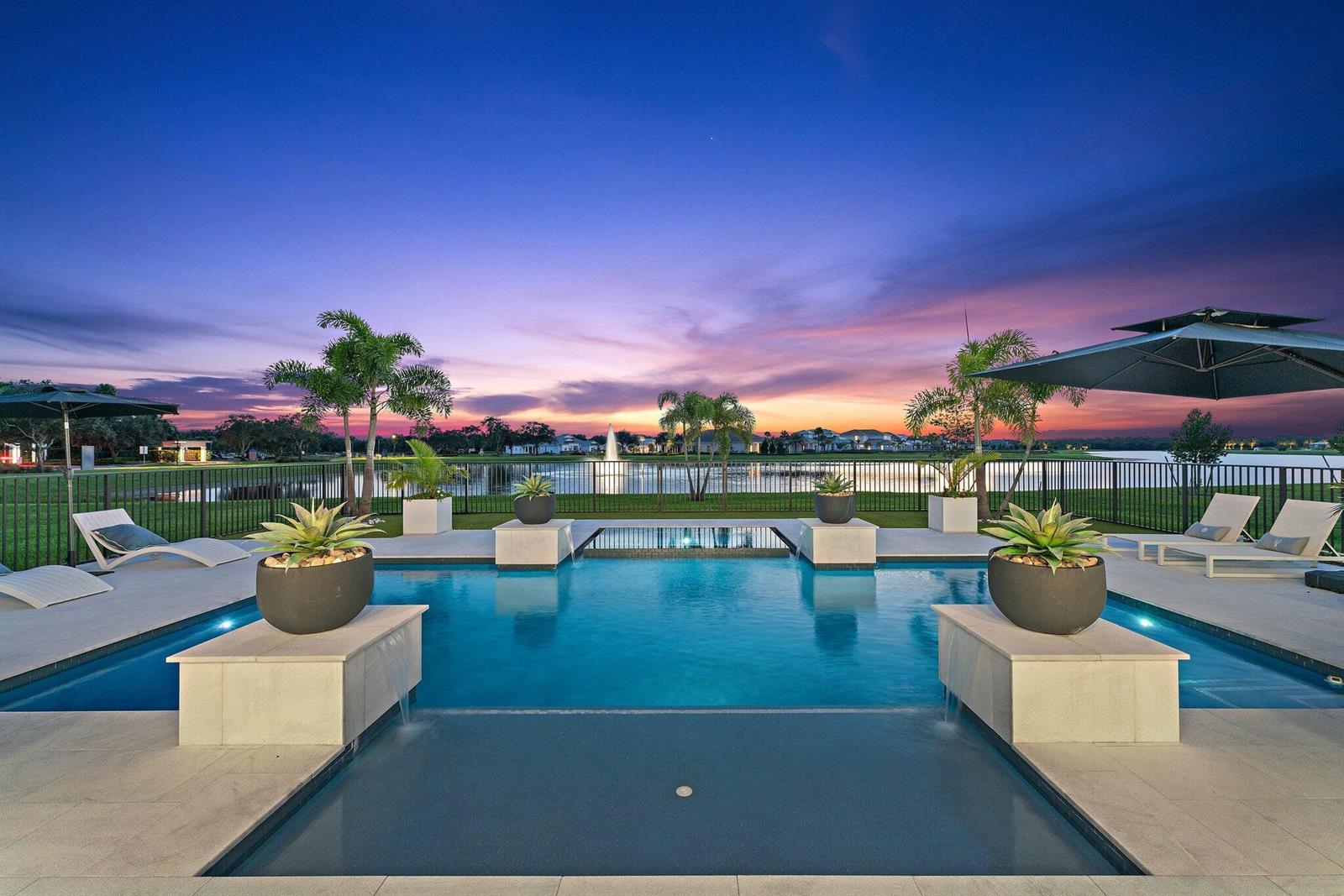 A swimming pool with water features and lounge chairs overlooks a lake at sunset — 20124 SE Bridgewater Drive, Jupiter FL 33458, JUPITER, FL