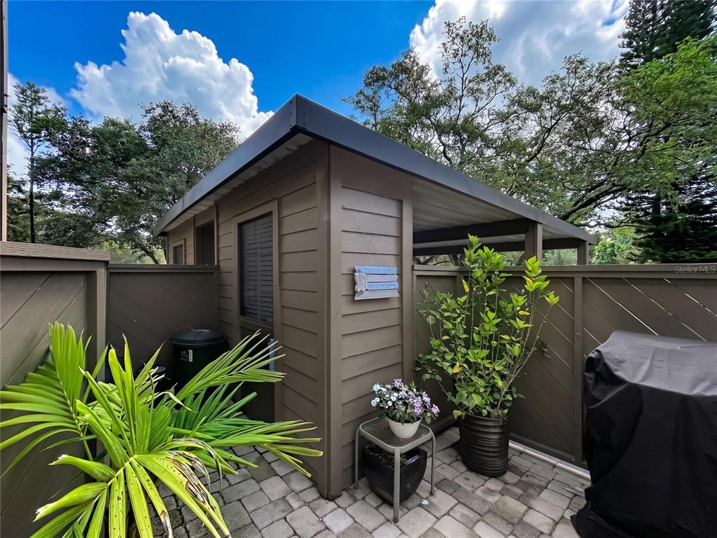 A brown shed with a covered patio area sits on a paved stone courtyard — 1006 LENNOX ROAD W UNIT 1006, PALM HARBOR, FL