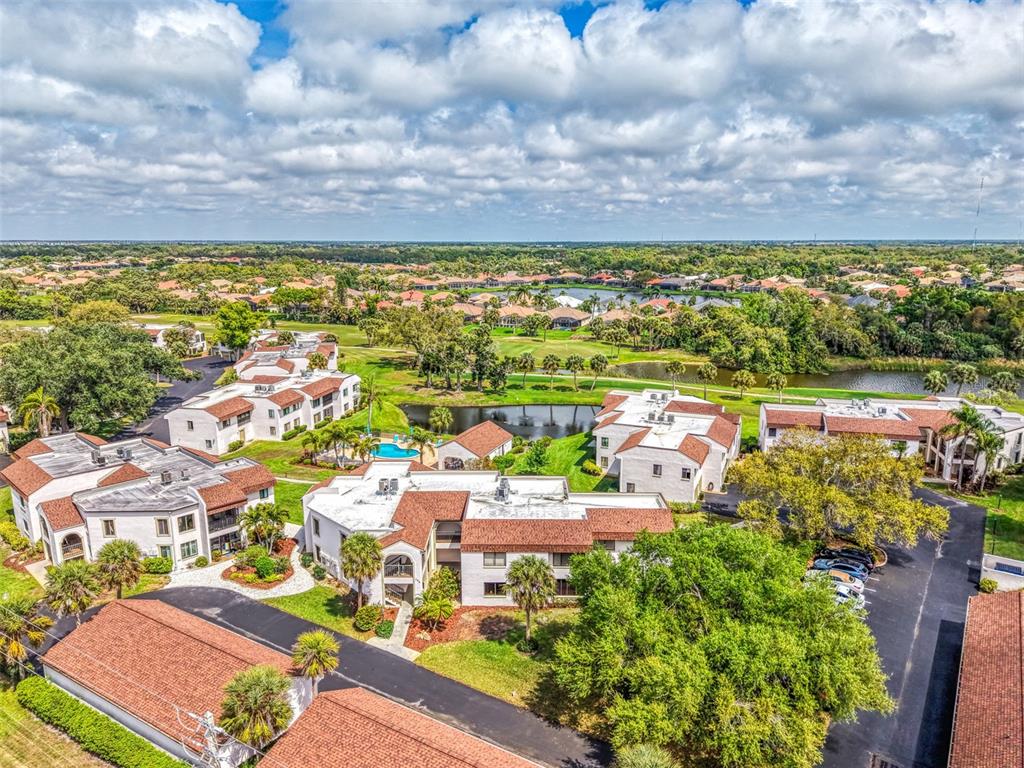 An aerial view shows multiple multi-story apartment buildings with red tile roofs near a pond — 796 CAPRI ISLES BOULEVARD UNIT 130, VENICE, FL