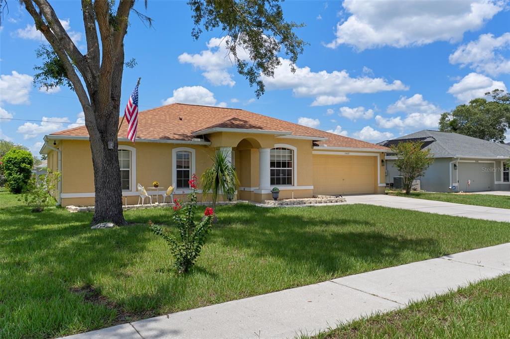 A single-story yellow house with a two-car garage, a large tree, and an American flag — 15437 DEER PATH DRIVE, BROOKSVILLE, FL