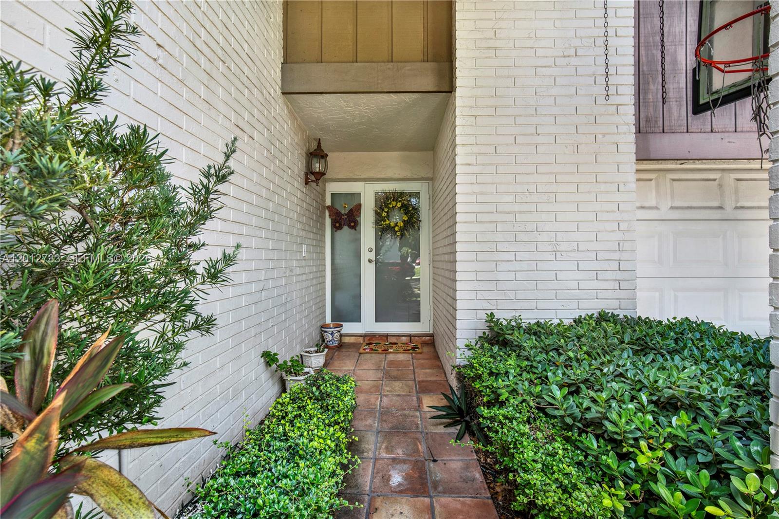 A white brick exterior entrance features a glass front door, tiled walkway, and green landscaping — 13455 SW 104th Ter, Miami FL 33186, MIAMI, FL