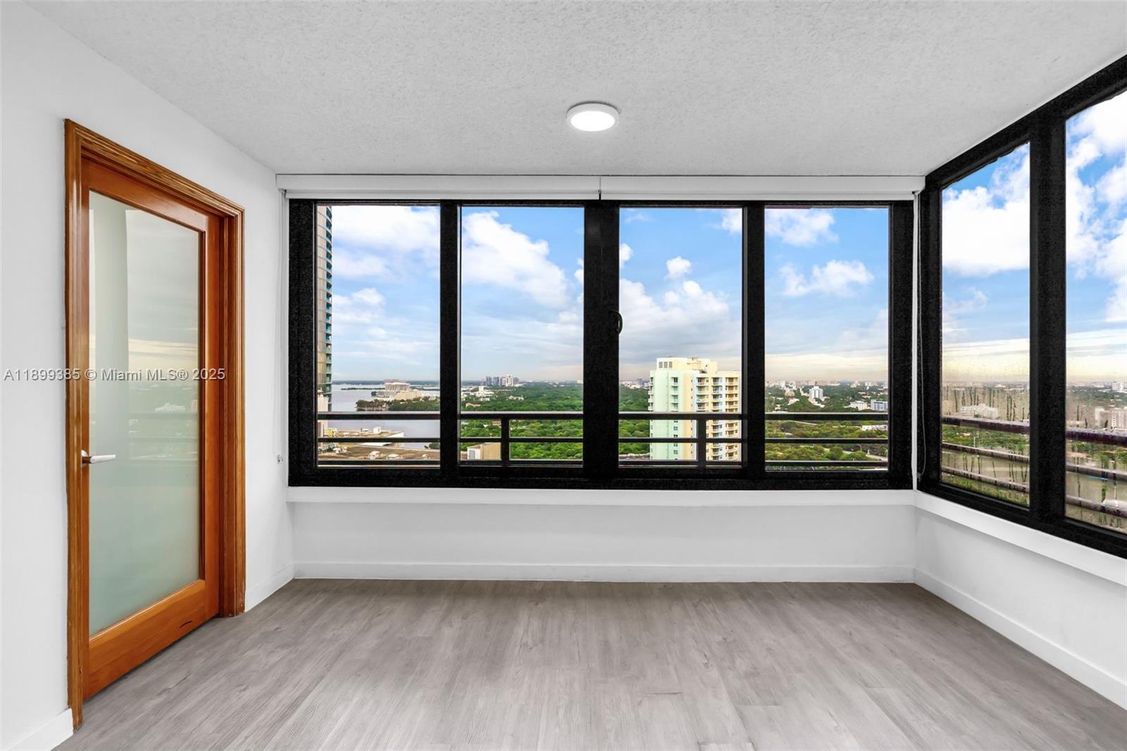 An empty room features light wood-look flooring, a wooden door, and large windows with city views — 2333 Brickell Ave, Miami FL 33129, MIAMI, FL