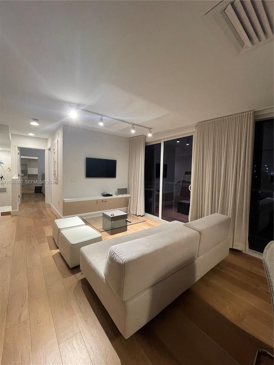 A modern living room features light wood flooring, a white sofa, and a wall-mounted television — 601 NE 27th St # 201, Miami FL 33137, MIAMI, FL