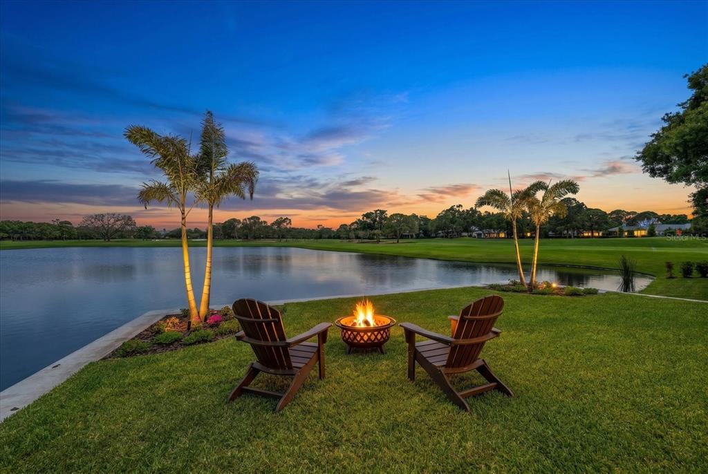 Two wooden chairs and a fire pit sit on a lawn overlooking a pond — 19 HIBISCUS ROAD, BELLEAIR, FL