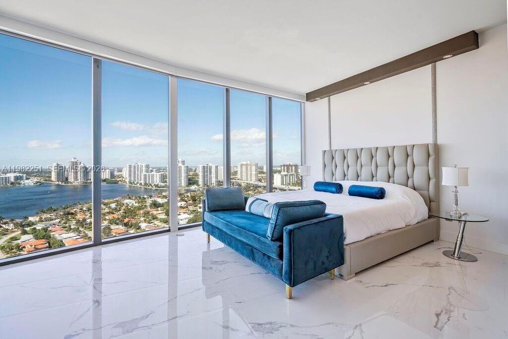 A modern bedroom features a tufted headboard, blue velvet bench, and floor-to-ceiling windows overlooking water — SUNNY ISLES BEACH, FL