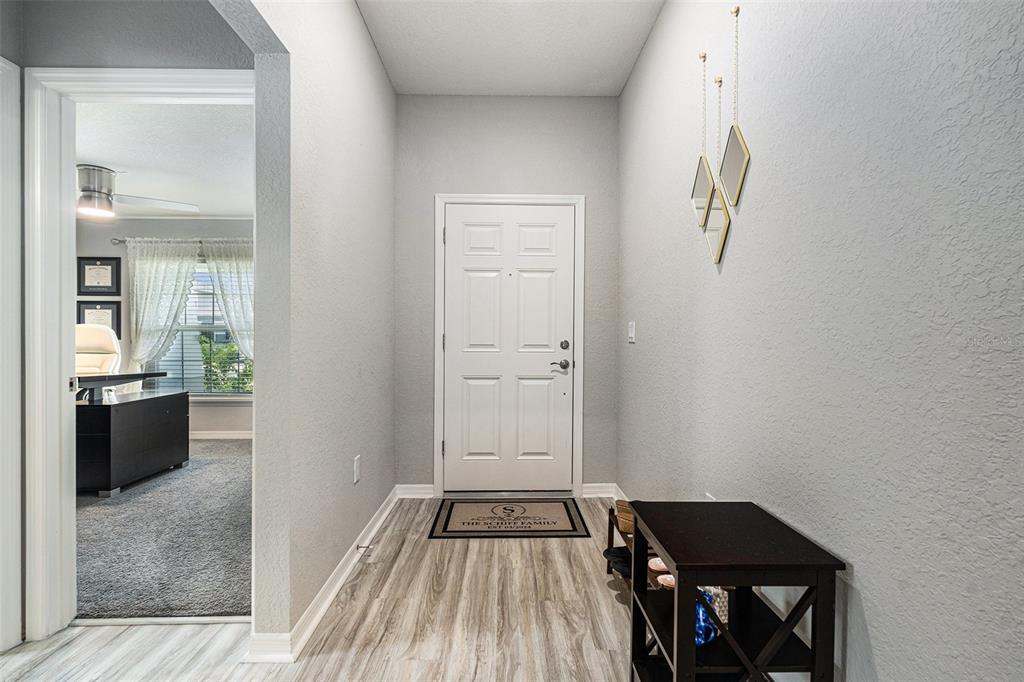 A bright entryway features light gray walls, wood-look flooring, a white front door, and decor — 6436 PEBBLEBROOKE WAY, ZEPHYRHILLS, FL