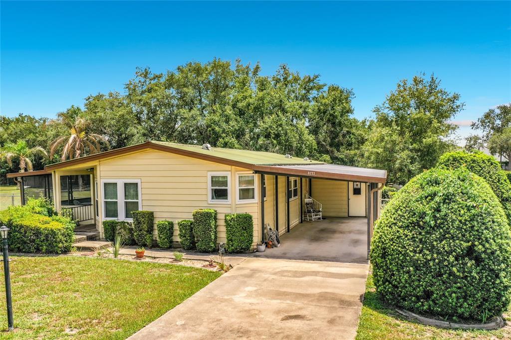 A single-story yellow house features a concrete driveway, a carport, and manicured landscaping — 8233 COUNTY ROAD 109B, LADY LAKE, FL