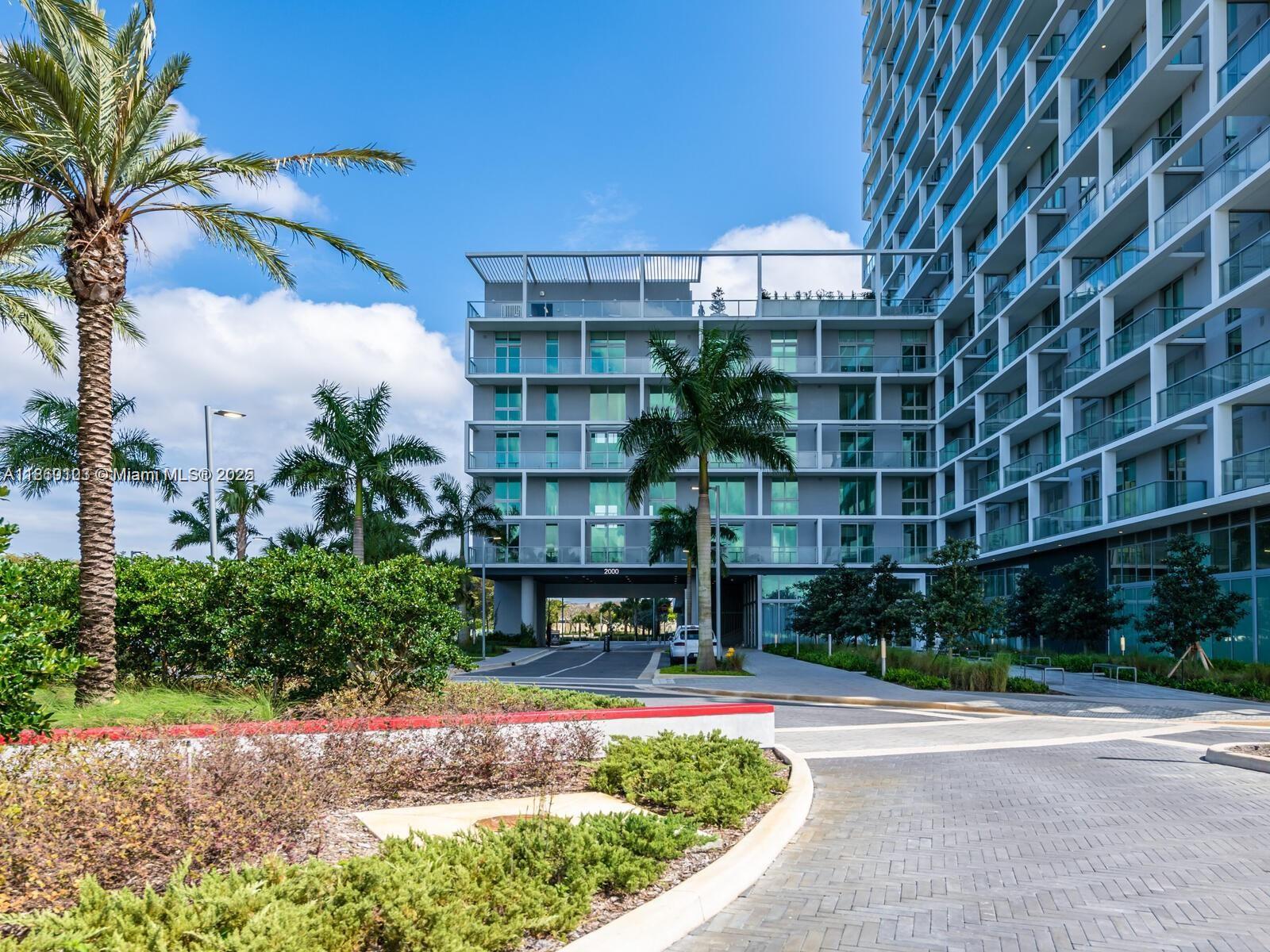 A modern multi-story apartment building with glass balconies stands behind a landscaped driveway and palms — 2000 Metropica Way # 2509, Sunrise FL 33323, SUNRISE, FL