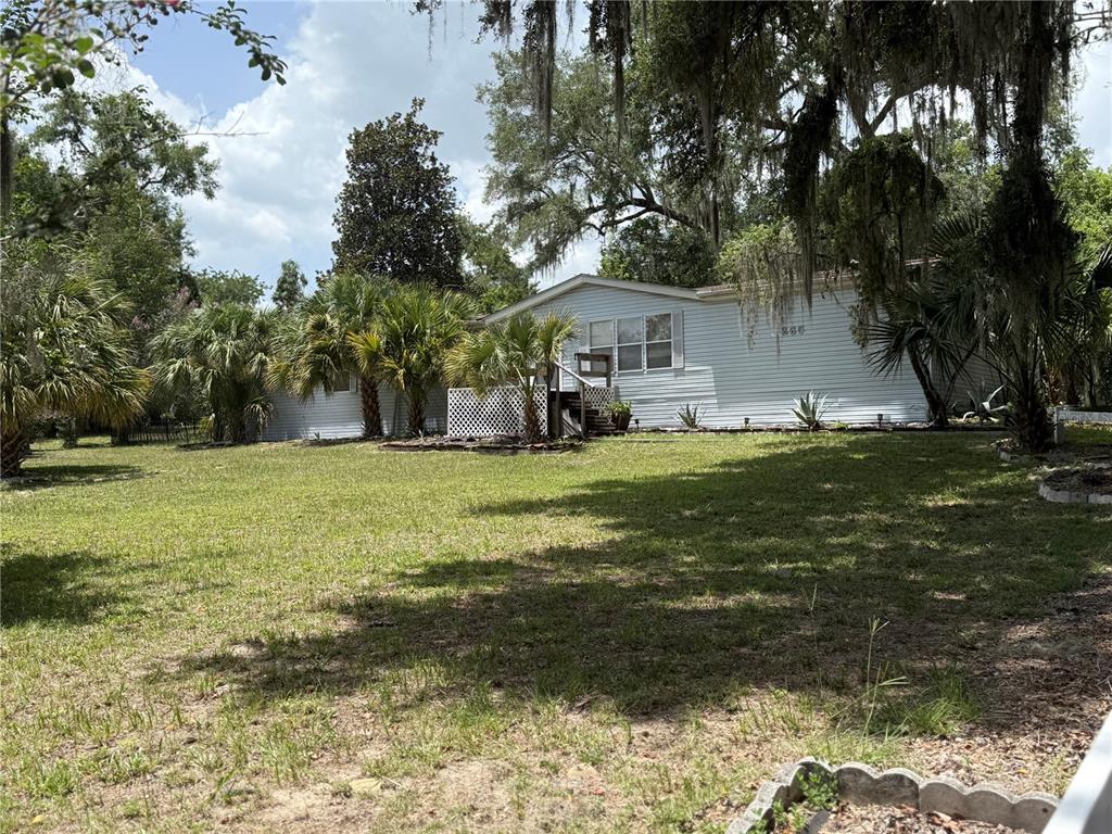A light-colored mobile home sits on a grassy lot surrounded by trees and palm plants — 265 W MCCLENDON ST, LADY LAKE, FL