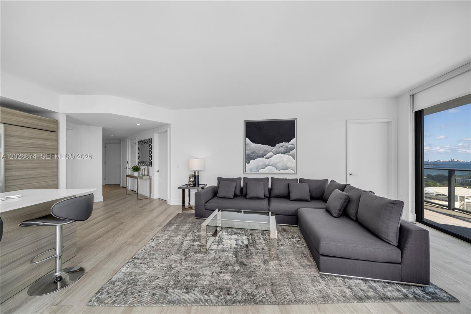 A modern living room features a gray sectional sofa, glass coffee table, and balcony view — 121 NE 34th St # 1704, Miami FL 33137, MIAMI, FL