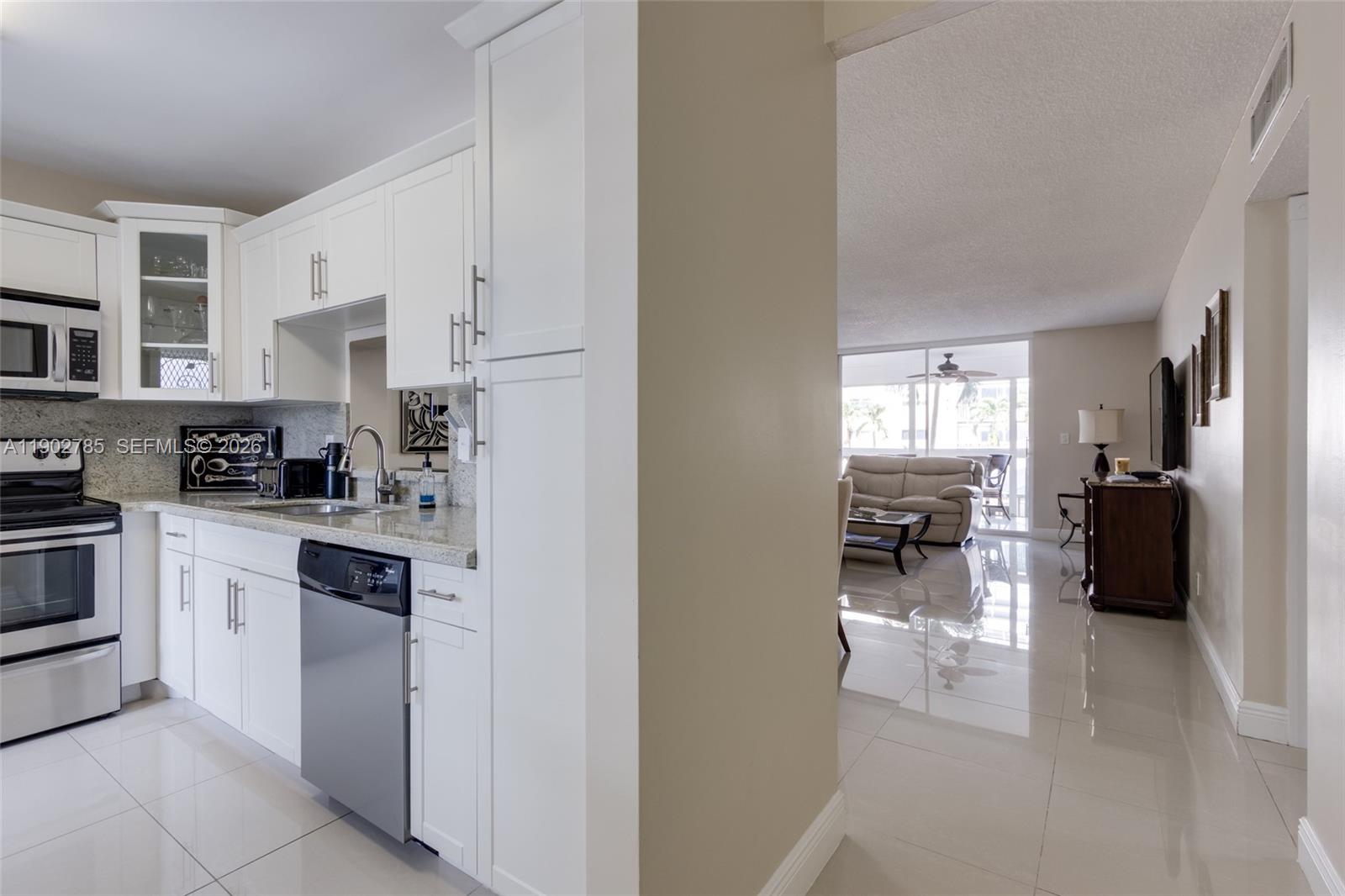 A white kitchen with stainless steel appliances opens into a living room with tiled floors — 420 NE 12th Ave # 208, Hallandale Beach FL 33009, HALLANDALE BEACH, FL