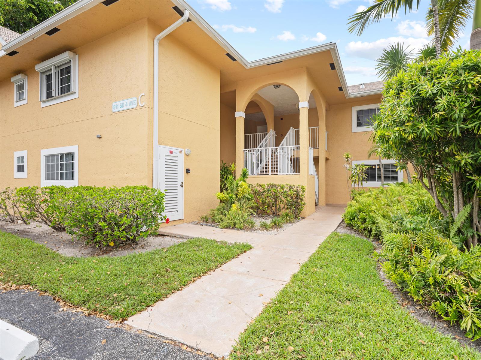 A two-story yellow stucco apartment building features an arched entryway with white stairs and landscaping — 811 SE 4th Avenue # C103, Dania Beach FL 33004, DANIA BEACH, FL