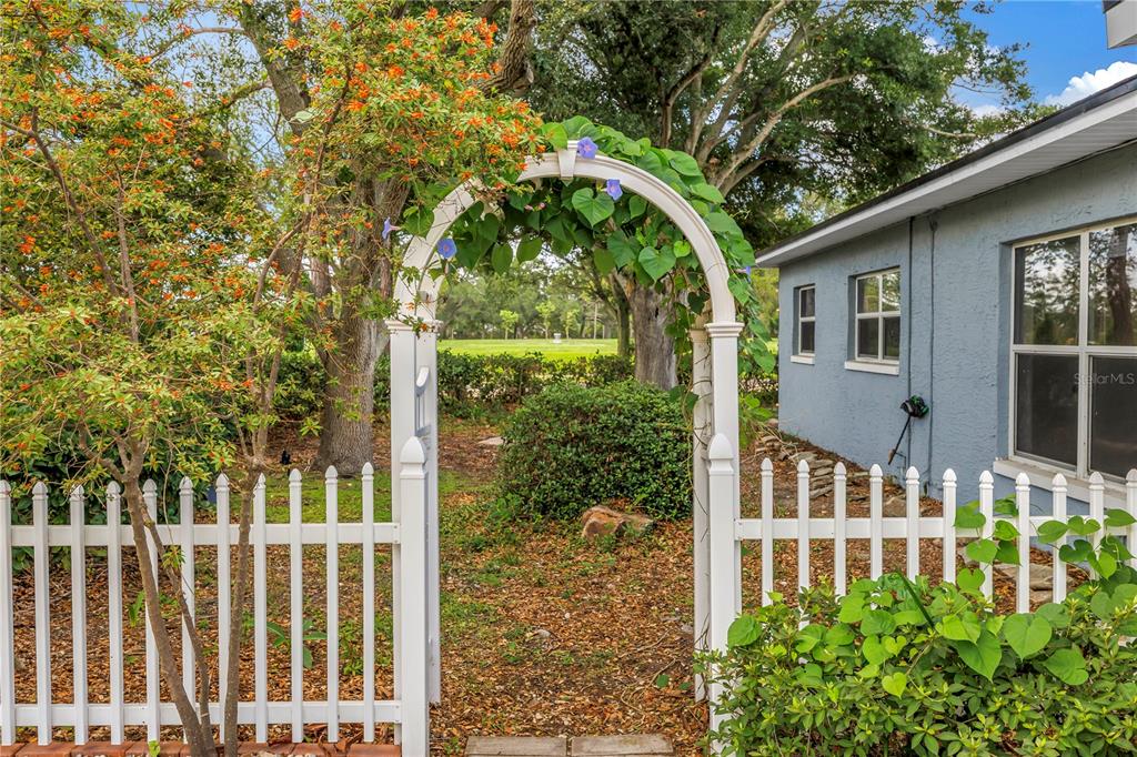 A white picket fence and arched gate stand beside a blue house with a garden — 7400 19TH AVENUE N, ST PETERSBURG, FL