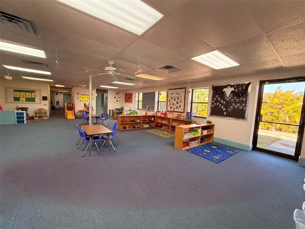 A spacious preschool classroom features blue carpeting, low shelving units, tables, and a glass door — 13610 LEARNING COURT, FORT MYERS, FL