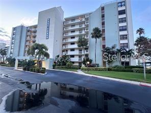 A multi-story apartment building with balconies and palm trees stands behind a paved parking lot — 300 WOODETTE DRIVE UNIT 303, DUNEDIN, FL
