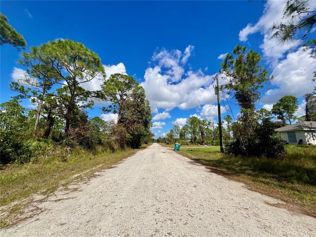 A gravel road stretches forward between pine trees under a bright blue sky with clouds — 12334 HINDLE AVENUE, PUNTA GORDA, FL