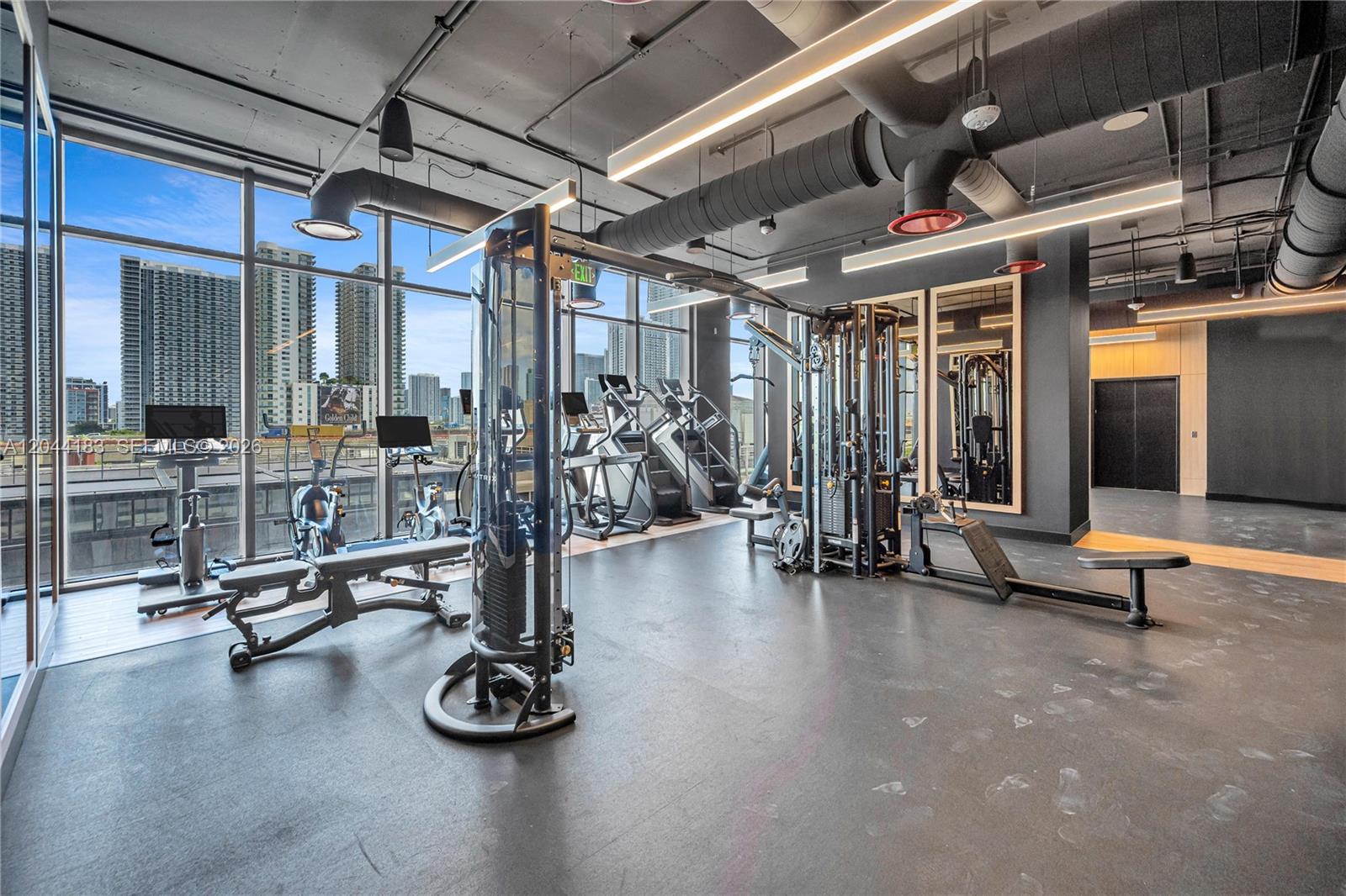 A modern gym features exercise machines, rubber flooring, and floor-to-ceiling windows overlooking a city skyline — 20 NE 11th, Miami FL 33132, MIAMI, FL