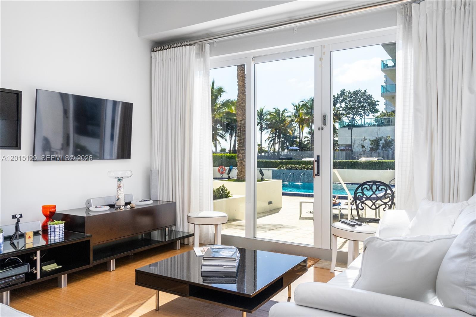 A living room with a television, dark furniture, and glass doors opening to a pool — 6917 Collins Ave # 103, Miami Beach FL 33141, MIAMI BEACH, FL