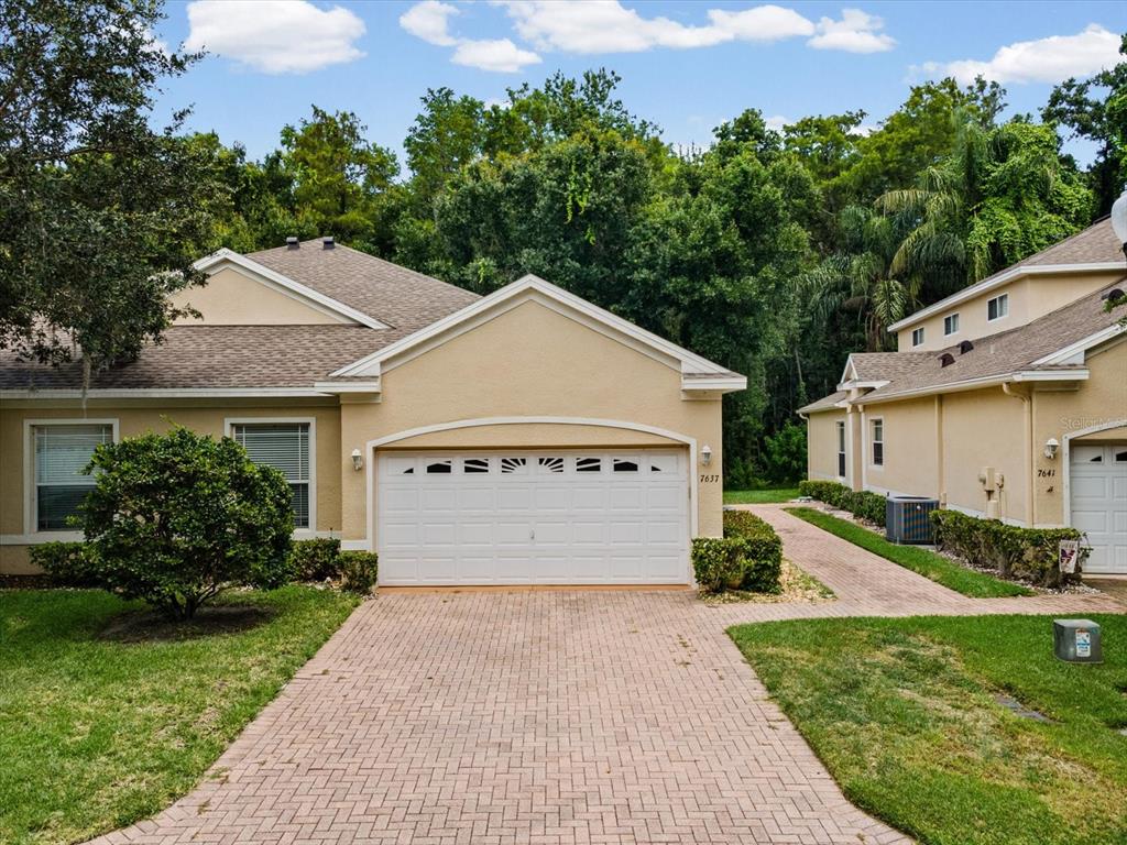 A tan stucco duplex features a two-car garage and a brick paver driveway — 7637 WIMPOLE DRIVE, NEW PORT RICHEY, FL