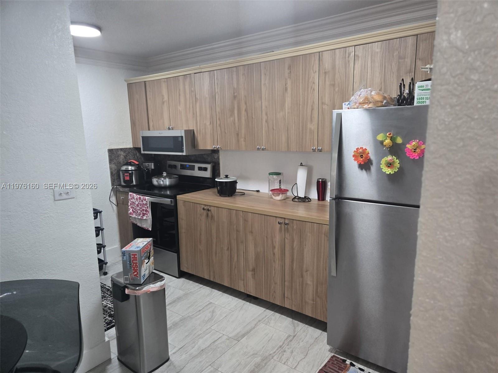 A kitchen features wood-grain cabinets, stainless steel appliances, and light-colored tile flooring — 1185 W 27th St, Hialeah FL 33010, HIALEAH, FL