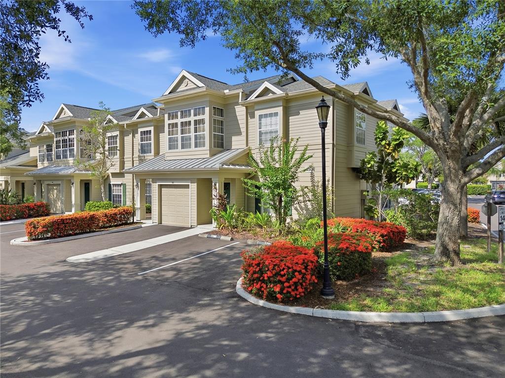 A two-story townhouse with tan siding, a garage, and red flowering shrubs in the foreground — 7780 PLANTATION CIRCLE, BRADENTON, FL