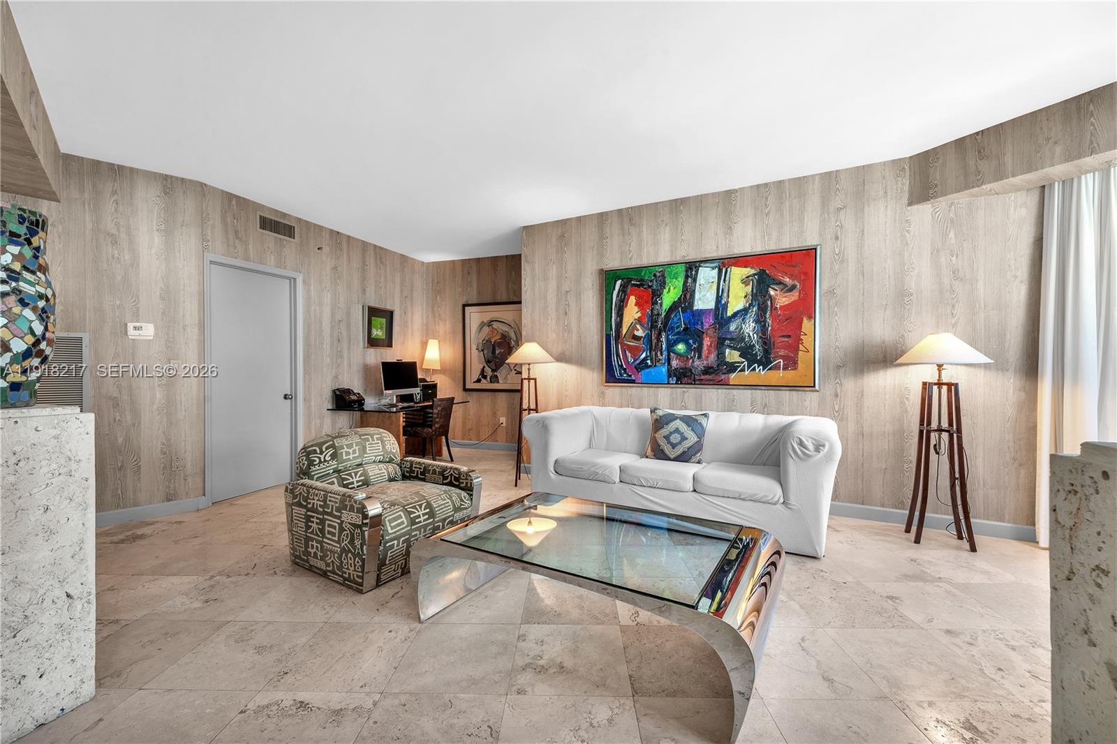 A living room features wood-paneled walls, a white sofa, a patterned armchair, and a glass-topped table — 300 S Pointe Dr # 906, Miami Beach FL 33139, MIAMI BEACH, FL