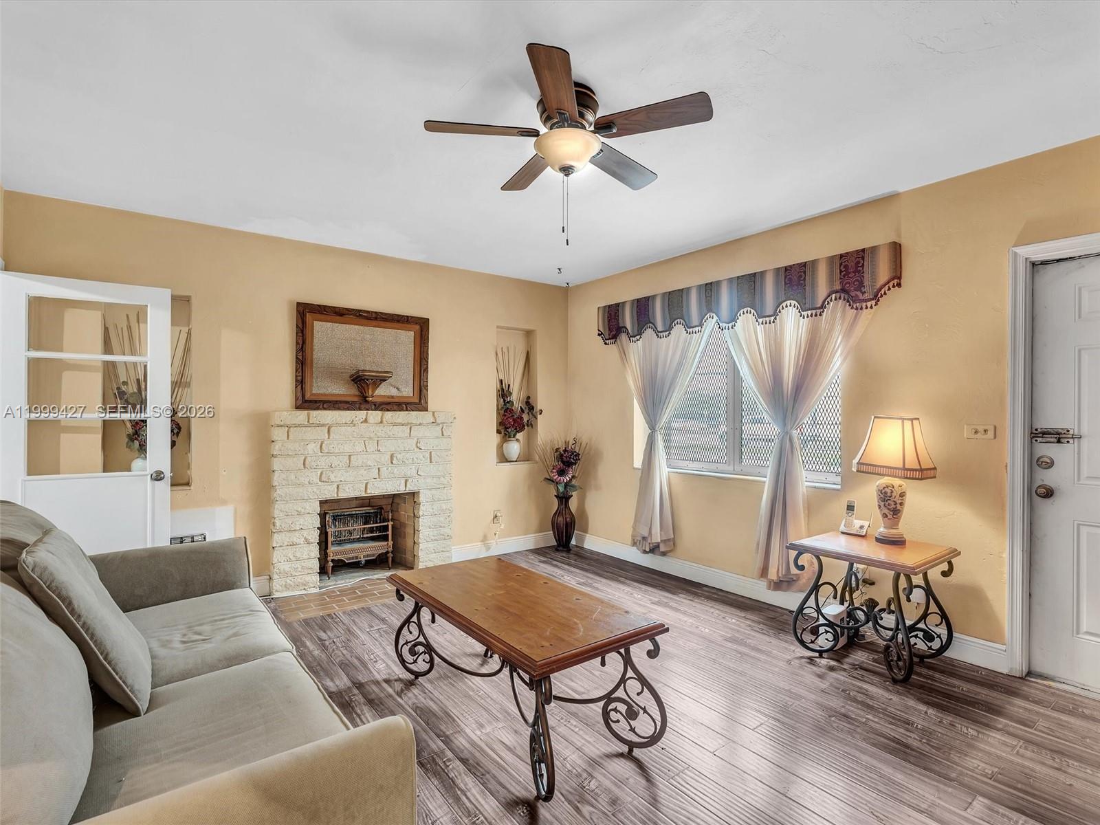 A living room features a brick fireplace, wood-look flooring, a ceiling fan, and beige walls — 10 SW 72nd Ave, Miami FL 33144, MIAMI, FL