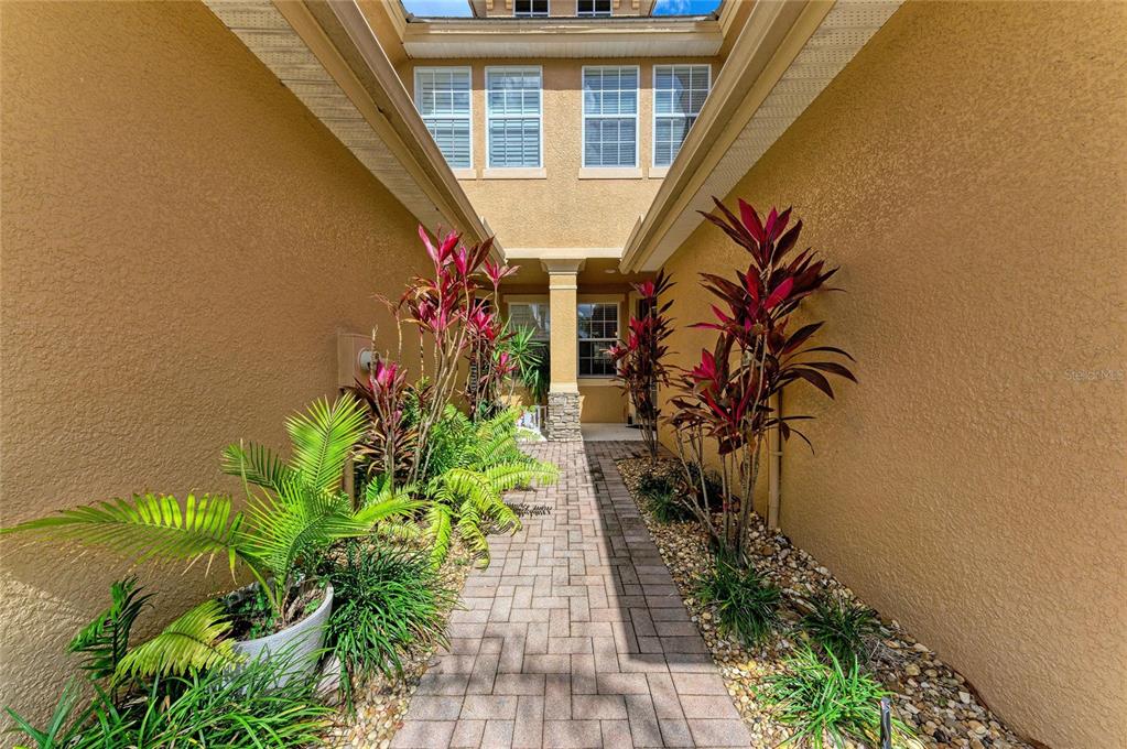 A paved walkway leads to a house entrance flanked by tropical plants and stone walls — 8160 VILLA GRANDE CT, SARASOTA, FL