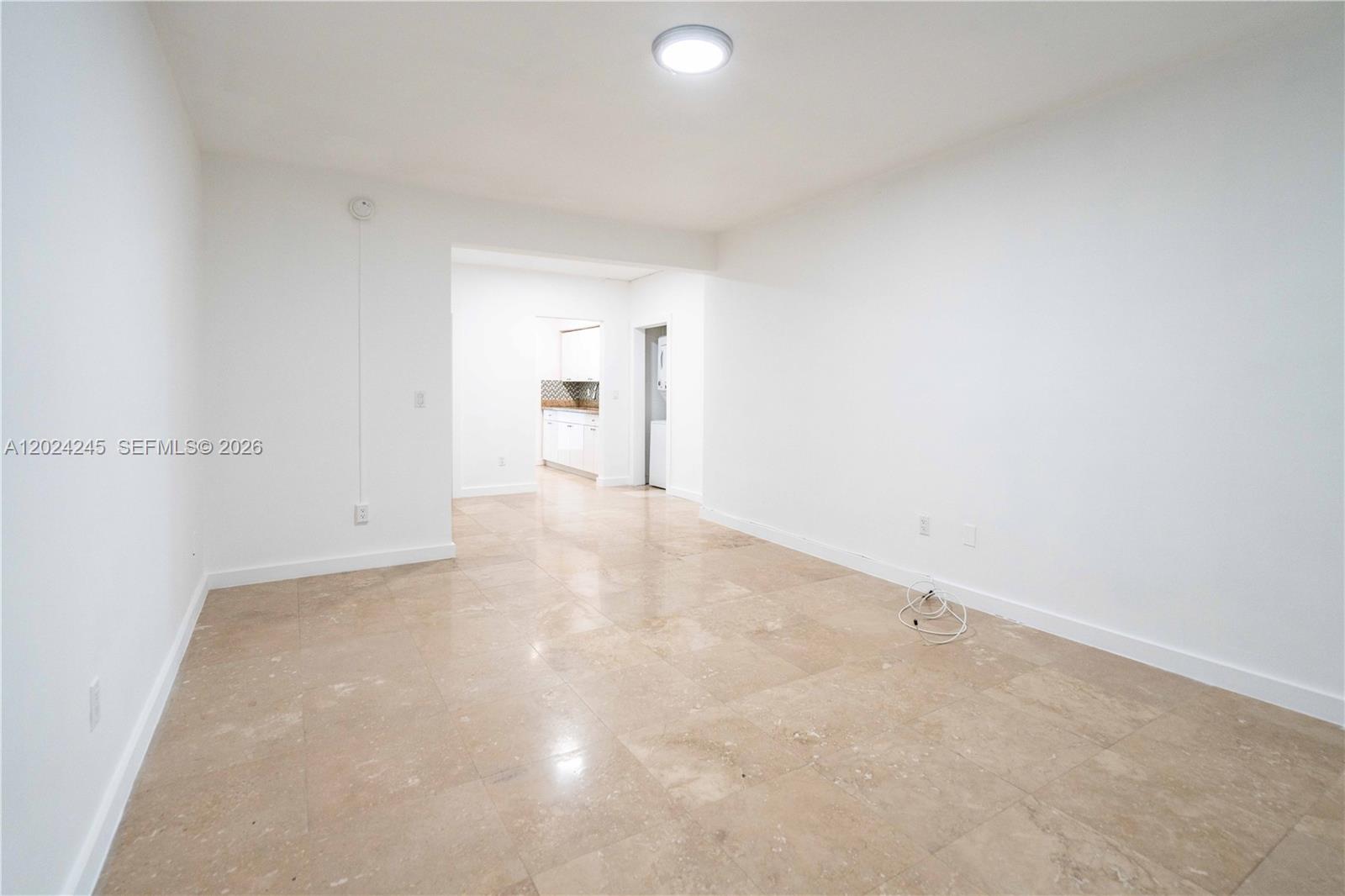 An empty room with white walls, beige tile flooring, and a view into a kitchen — 1120 Euclid Ave # 6, Miami Beach FL 33139, MIAMI BEACH, FL