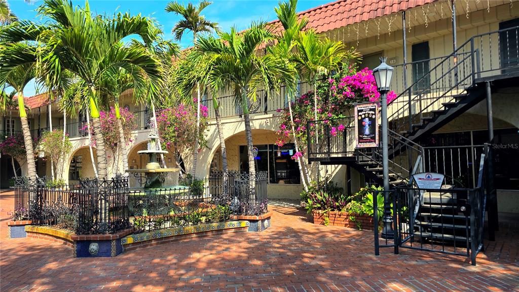 A brick courtyard features a tiered fountain, palm trees, and a two-story building with balconies — 530 US-41-BYP UNIT 1A, VENICE, FL